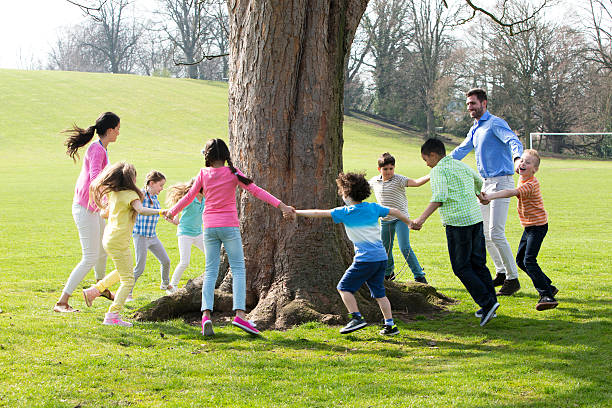 A group of children with two adults holding hands and dance around the trunk of a tree.